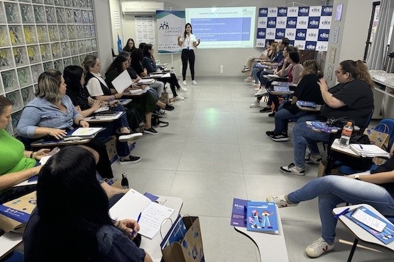 A imagem mostra uma sala de aula ou reunião, onde várias pessoas estão sentadas em cadeiras dispostas em um formato de U. Elas estão prestando atenção em uma mulher que está em pé na frente da sala, aparentemente dando uma apresentação. A mulher está de pé ao lado de uma tela de projeção que exibe slides com texto. As pessoas na sala estão com cadernos e materiais de anotação, e algumas têm sacolas de papel ao lado. As paredes têm uma decoração com logotipos repetidos, e há uma parede de blocos de vidro à esquerda. A atmosfera parece ser de um ambiente educacional ou de treinamento.
