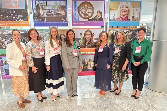 A imagem mostra um grupo de sete mulheres posando para uma foto em frente a um painel de fotos e cartazes. Todas estão vestidas de forma elegante, com roupas formais ou semi-formais. O painel atrás delas exibe várias imagens, incluindo uma com o logotipo das Nações Unidas e outras com retratos de pessoas em eventos ou reuniões. As mulheres estão sorrindo e parecem estar participando de um evento ou conferência. O ambiente parece ser um espaço interno, possivelmente um saguão ou área de exposição.