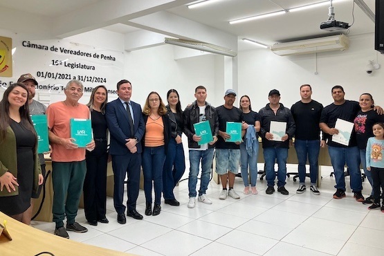 A imagem mostra um grupo de pessoas posando para uma foto em um ambiente interno, que parece ser uma sala de reuniões ou conferências. Ao fundo, há uma parede com o texto "Câmara de Vereadores de Penha" e informações sobre a legislatura de 2021 a 2024. O grupo é composto por 15 pessoas, incluindo homens e mulheres, que estão em pé lado a lado. Algumas pessoas estão segurando pastas verdes com a inscrição "Lar Legal". No canto direito da imagem, há uma mulher abraçando uma criança. O ambiente é bem iluminado, com piso branco e teto com luminárias.