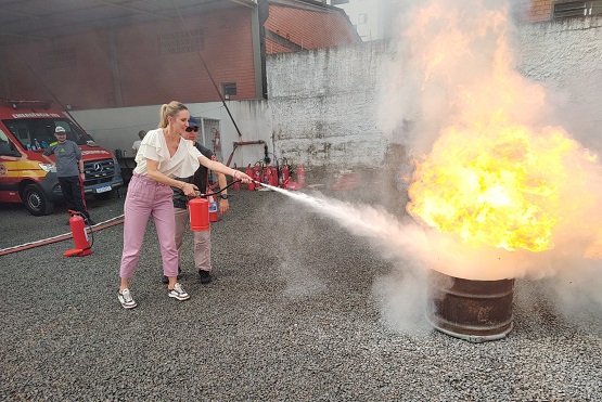 A imagem mostra uma mulher usando um extintor de incêndio para apagar um fogo em um tambor de metal. Ela está vestindo uma blusa branca e calça rosa, e parece estar em um ambiente ao ar livre com chão de cascalho. Ao fundo, há um homem observando a cena, e um veículo de emergência vermelho estacionado. O fogo no tambor está bastante intenso, com chamas altas e fumaça. A mulher está acompanhada por outra pessoa que parece estar orientando-a no uso do extintor.