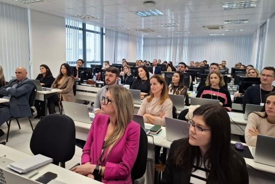 A imagem que você enviou mostra uma sala de aula ou conferência com várias pessoas sentadas, todas focadas em uma apresentação ou aula que está acontecendo à frente. As pessoas estão em mesas brancas, com laptops abertos em frente a elas. A maioria dos participantes parece atenta e concentrada, alguns fazendo anotações. Há uma variedade de pessoas, tanto homens quanto mulheres, vestidas de maneira formal e casual. O ambiente é moderno, com janelas grandes ao fundo, deixando entrar luz natural, e há persianas brancas cobrindo as janelas. O teto é branco com luminárias embutidas, iluminando bem a sala. A atmosfera parece ser profissional, talvez um treinamento ou workshop empresarial.