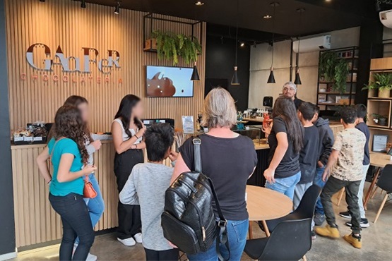 A imagem mostra o interior de uma cafeteria moderna e bem iluminada. Há um grupo de pessoas, incluindo adultos e crianças, em pé em frente ao balcão de atendimento. O balcão é feito de madeira clara com ripas verticais, e acima dele está escrito "CAFÉ" em letras grandes. Há também a palavra "CHOCOLATERIA" em letras menores logo abaixo. No balcão, há algumas plantas decorativas e um monitor que parece exibir uma imagem de chocolate. As pessoas estão conversando e algumas parecem estar fazendo pedidos. O ambiente é decorado com plantas suspensas e há mesas e cadeiras ao redor para os clientes.