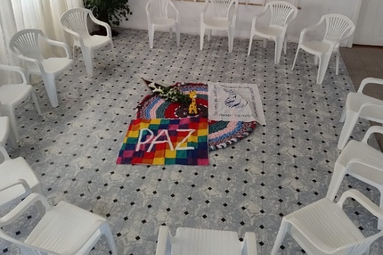 A imagem mostra uma sala com várias cadeiras de plástico branco dispostas em círculo. No centro do círculo, há um arranjo no chão composto por um tapete colorido, flores e dois panos. Um dos panos é colorido com quadrados em várias cores e a palavra "PAZ" escrita em branco. O outro pano é branco com um desenho de uma pomba e as palavras ilegíveis. A sala tem um piso claro com um padrão geométrico e há uma planta no canto.