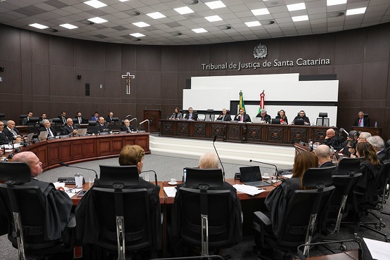 A imagem mostra uma sessão em andamento no Tribunal de Justiça de Santa Catarina. A sala é ampla, com um grande número de pessoas sentadas em cadeiras dispostas em formato semicircular. Na parte central, há uma bancada elevada onde estão sentados vários juízes ou desembargadores, todos vestidos com trajes formais. Atrás deles, na parede, está escrito "Tribunal de Justiça de Santa Catarina" e há um brasão acima do texto. À esquerda, há um crucifixo fixado na parede. A bandeira do Brasil e a bandeira do estado de Santa Catarina estão posicionadas atrás da bancada central. A sala tem um teto com iluminação embutida e um piso acarpetado.