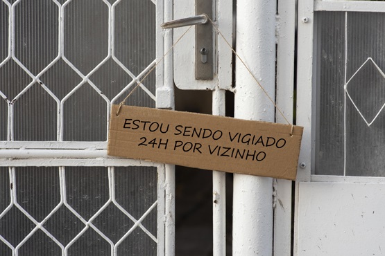 A imagem mostra uma porta de metal com uma grade de proteção. Na porta, há uma placa de papelão pendurada por um barbante. A placa tem a seguinte mensagem escrita: "ESTOU SENDO VIGIADO 24H POR VIZINHO".