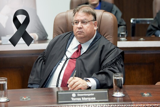 A imagem mostra um homem sentado em uma cadeira de couro marrom, vestindo uma toga preta sobre uma camisa branca e gravata vermelha. Ele está em um ambiente que parece ser uma sala de tribunal ou uma sessão formal, com outras pessoas ao fundo. Na mesa à sua frente, há um copo de água e uma placa com o nome "Desembargador Torres Marques". No canto superior esquerdo da imagem, há um laço preto, que geralmente simboliza luto.