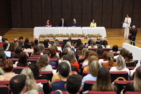 A imagem mostra um auditório com várias pessoas sentadas, todas voltadas para um palco onde há uma mesa comprida coberta com uma toalha branca. Atrás da mesa, estão sentadas quatro pessoas, três homens e uma mulher, e há uma quinta pessoa em pé, falando em um púlpito à direita. A mesa está decorada com arranjos de flores na frente. O auditório está cheio, com pessoas de diferentes idades e estilos de cabelo, e parece ser um evento formal ou uma conferência.