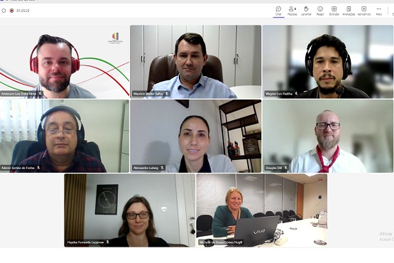 A imagem mostra uma captura de tela de uma videoconferência com oito participantes, cada um em sua própria janela de vídeo.  1. No canto superior esquerdo, há um homem com fones de ouvido vermelhos e uma camisa cinza.  2. Ao lado dele, está um homem vestindo uma camisa azul clara e sentado em uma cadeira de escritório. 3. No canto superior direito, está um homem usando fones de ouvido pretos e uma camisa escura. 4. No meio à esquerda, está um homem, usando óculos e fones de ouvido, com uma camisa xadrez. 5. No centro, está uma mulher com cabelo preso e uma blusa clara. 6. No meio à direita, está um homem usando óculos e uma camisa branca com gravata vermelha. 7. No canto inferior esquerdo, está uma mulher com cabelo solto e óculos, em frente a uma parede com um quadro. 8. No canto inferior direito, está uma mulher loira, sorrindo, com um laptop Vaio à sua frente.  A interface da videoconferência mostra ícones no topo para controle de áudio, vídeo e outras funções.