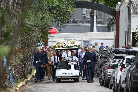 A imagem mostra um cortejo fúnebre em um cemitério. No centro, há um carrinho de golfe branco com a inscrição "Jardim da Luz" na frente, transportando um caixão coberto por flores brancas e amarelas. O carrinho é escoltado por homens uniformizados, possivelmente militares, que caminham ao lado. Há várias pessoas seguindo o cortejo, algumas vestidas de terno. O caminho é ladeado por árvores à esquerda e carros estacionados à direita. Ao fundo, é possível ver um portão e algumas construções. A atmosfera é solene e respeitosa.