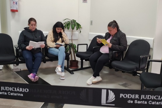 A imagem mostra três mulheres sentadas em uma sala de espera. Elas estão em cadeiras pretas, lado a lado. A mulher à esquerda está usando um moletom escuro e tênis roxos, e está lendo um livro. A mulher no meio está vestindo uma jaqueta bege e calça jeans, e está lendo um livro. A mulher à direita está usando uma jaqueta escura e calça preta, e está lendo um livro ou caderno amarelo. Há uma planta em um vaso no chão, atrás delas. Na frente, há uma fita preta com o texto "Poder Judiciário de Santa Catarina". Ao fundo, na parede, há um extintor de incêndio.