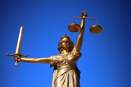A imagem mostra uma estátua dourada da deusa da justiça, conhecida como "Justiça Cega". Ela está segurando uma balança na mão esquerda e uma espada na mão direita. A estátua está vendada, simbolizando imparcialidade. O fundo é um céu azul claro.