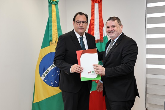A imagem mostra dois homens de terno e gravata posando para uma foto. Eles estão em um ambiente interno, provavelmente um escritório ou sala de reuniões. O homem à esquerda está segurando uma pasta com as cores vermelha e verde, que tem o logotipo do Ministério Público de Santa Catarina (MPSC) na frente. Atrás deles, há duas bandeiras: a bandeira do Brasil à esquerda e a bandeira do estado de Santa Catarina à direita. Ambos os homens estão sorrindo para a câmera.