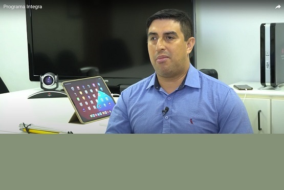 A imagem mostra um homem de pele clara e cabelo curto e escuro, vestindo uma camisa azul clara. Ele está sentado em uma sala de escritório, com um tablet apoiado em um suporte ao seu lado, mostrando a tela inicial com vários ícones de aplicativos. Atrás dele, há uma grande televisão ou monitor e uma câmera de videoconferência. À direita, há um computador de mesa ou servidor. No canto superior esquerdo da imagem, está escrito "Programa Integra".