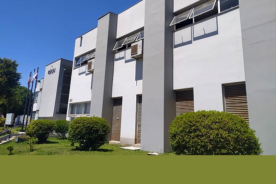 A imagem mostra um edifício de dois andares com uma fachada moderna e de cor clara. Há várias janelas com persianas e algumas delas estão abertas. No lado esquerdo da imagem, há bandeiras hasteadas, sugerindo que o local pode ser uma instituição pública ou governamental. Na frente do edifício, há arbustos bem cuidados e uma área gramada. O céu está azul, indicando que é um dia ensolarado.