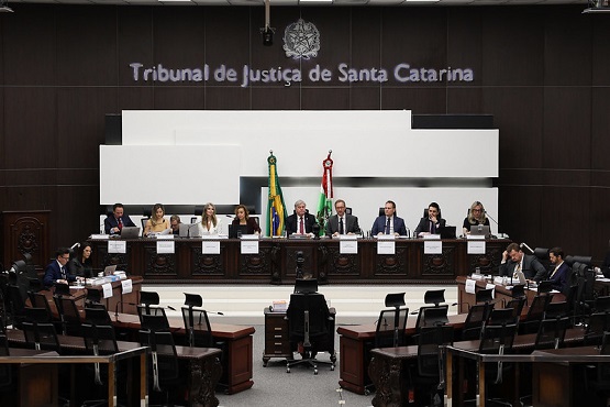 A imagem mostra uma sessão no Tribunal de Justiça de Santa Catarina. Há uma mesa comprida onde estão sentadas várias pessoas, provavelmente juízes ou autoridades judiciais. Atrás deles, há duas bandeiras, uma do Brasil e outra de Santa Catarina. Acima, na parede, está escrito "Tribunal de Justiça de Santa Catarina". A sala é grande, com várias cadeiras e mesas dispostas em um formato que lembra um auditório ou plenário. A decoração é sóbria, com tons escuros predominando.