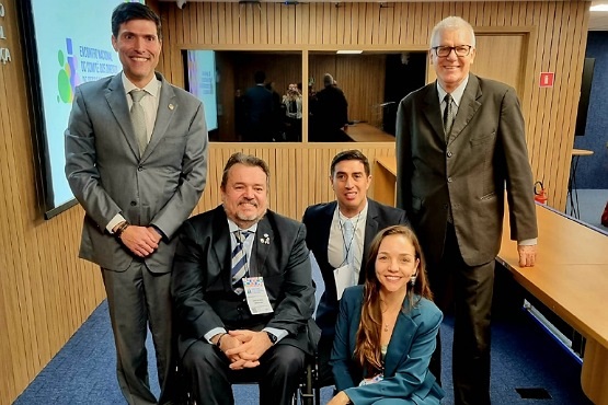 A imagem mostra cinco pessoas em um ambiente interno, provavelmente uma sala de conferências ou reunião. Dois homens estão em pé e dois homens e uma mulher estão sentados na frente. Todos estão vestidos formalmente com ternos.  O homem à esquerda está sorrindo e usando um terno cinza claro. Ao lado dele, à direita, está um homem de terno escuro, sentado em uma cadeira de rodas, também sorrindo. Ao lado dele, está uma mulher ajoelhada, sorrindo, usando um terno azul. Atrás dela, está um homem de terno escuro, também sorrindo. À direita, está outro homem de terno escuro, com óculos, sorrindo. No fundo, há uma parede de madeira com um espelho grande, refletindo parte das pessoas e do ambiente. À esquerda, há uma tela de projeção com um logotipo colorido e algumas palavras, mas não é possível ler o texto completo. A sala tem um carpete azul e uma mesa de madeira à direita.