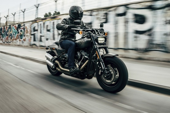 A imagem mostra uma pessoa pilotando uma motocicleta em alta velocidade. A motocicleta é preta e o piloto está usando um capacete e roupas escuras. Ao fundo, há um muro com grafites e arame farpado no topo. A imagem parece capturar um momento de movimento rápido, com um efeito de desfoque no fundo.