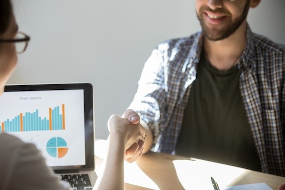 A imagem mostra duas pessoas apertando as mãos. Uma delas está parcialmente visível, usando uma camisa xadrez e óculos, e parece estar sorrindo. Ao fundo, há um monitor de computador exibindo um gráfico de barras e um gráfico de pizza.