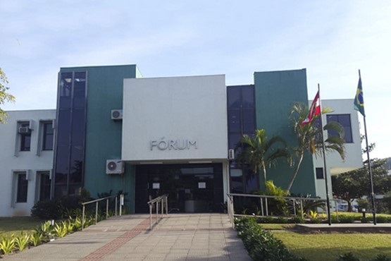 A imagem mostra a fachada de um prédio identificado como "FÓRUM". O edifício tem um design moderno, com linhas retas e é predominantemente branco com detalhes em verde. Há janelas verticais de vidro escuro em ambos os lados da entrada principal. Na frente do prédio, há uma rampa de acesso e um caminho pavimentado que leva até a entrada. À direita, há duas bandeiras hasteadas, uma do Brasil e outra de Santa Catarina. Há também algumas plantas e palmeiras ao redor, contribuindo para a paisagem do local. O céu está claro, indicando que a foto foi tirada durante o dia.