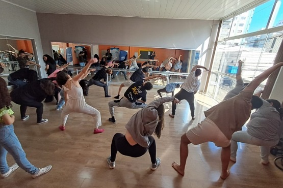 A imagem mostra um grupo de pessoas em uma sala de dança, realizando um alongamento ou exercício físico. Elas estão posicionadas em fileiras, todas inclinadas para o lado direito com um braço estendido acima da cabeça. A sala tem piso de madeira clara e uma parede com espelhos, refletindo os participantes. Há uma grande janela à direita, permitindo a entrada de luz natural. As pessoas estão vestidas de maneira casual, com roupas confortáveis para a atividade física.