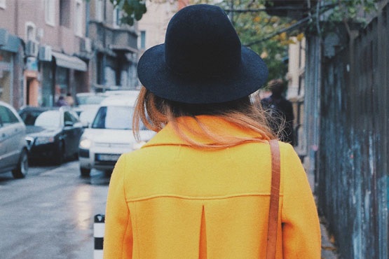 A imagem mostra uma pessoa de costas caminhando em uma rua. Ela está usando um casaco amarelo vibrante e um chapéu preto. O cabelo dela é castanho claro e está solto. Ao fundo, há carros estacionados e prédios de aparência urbana. A cena parece ser em um dia nublado ou com pouca luz natural.