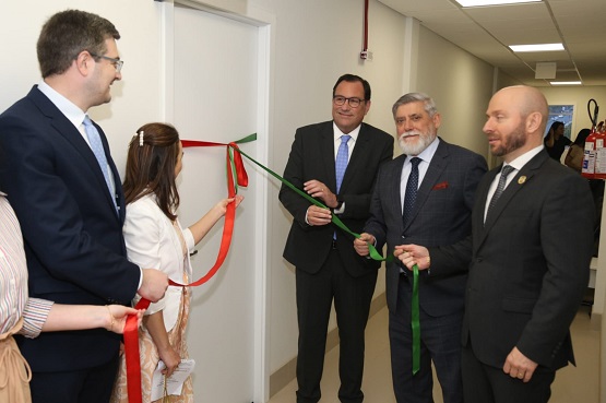 A imagem mostra seis pessoas em um evento de inauguração, onde estão cortando uma fita vermelha e verde. As pessoas estão em um corredor, em frente a uma porta branca. Da esquerda para a direita, há um braço de uma mulher com que veste uma blusa de malha, um homem de óculos e terno escuro, uma mulher de cabelos castanhos e vestido claro, um homem de óculos e terno escuro com gravata azul, um homem de barba grisalha e terno escuro com gravata azul e lenço vermelho no bolso, e um homem careca com barba e terno escuro. Todos parecem estar sorrindo e participando do corte da fita. Ao fundo, o corredor é iluminado e há um extintor de incêndio na parede.