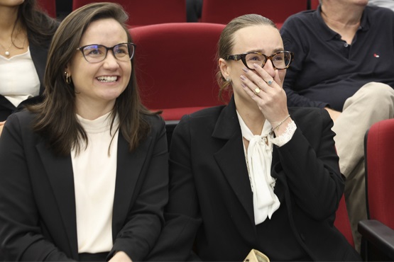 A imagem mostra duas mulheres sentadas lado a lado em cadeiras vermelhas, aparentemente em um auditório. Ambas estão usando óculos e vestem roupas formais, com blazers pretos e blusas claras. A mulher à esquerda está sorrindo amplamente, enquanto a mulher à direita está com a mão esquerda cobrindo parcialmente a boca, como se estivesse surpresa ou chorando. Ao fundo, há outras pessoas sentadas, mas elas estão fora de foco.