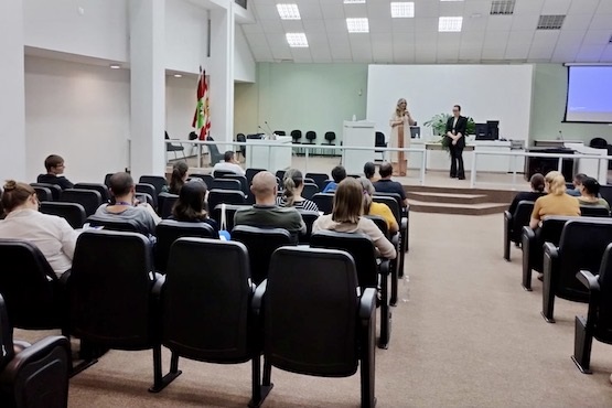 A imagem mostra uma sala de sessões ou auditório com várias fileiras de cadeiras pretas, onde algumas pessoas estão sentadas, voltadas para a frente da sala. Na frente, há um palco elevado com duas mulheres em pé, que parecem estar se apresentando ou discursando. Atrás deles, há uma mesa comprida com cadeiras e equipamentos eletrônicos, como computadores. Na parede do fundo, há uma tela de projeção branca e, à direita, uma tela azul que parece ser parte de um projetor. No lado esquerdo da imagem, há bandeiras em um suporte. A sala tem um teto com painéis de luz embutidos.