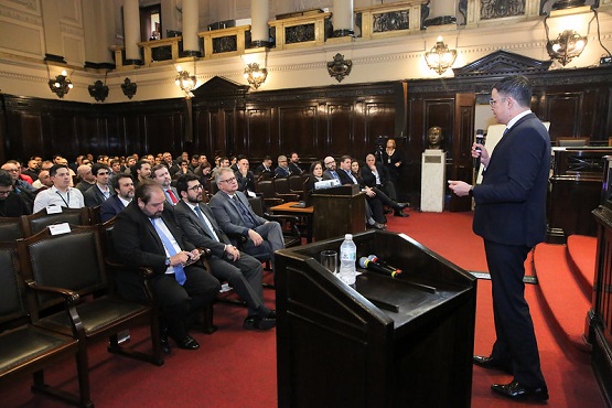 A imagem mostra uma sala de conferências ou auditório com várias pessoas sentadas, a maioria delas usando ternos, o que sugere um evento formal ou profissional. À direita, um homem de terno está em pé, segurando um microfone, aparentemente fazendo uma apresentação ou discurso. O ambiente é elegante, com paredes de madeira escura e detalhes arquitetônicos clássicos, como colunas e luminárias antigas. Há uma mesa com um microfone e uma garrafa de água em frente ao homem que está falando. As pessoas na plateia estão prestando atenção ao orador.