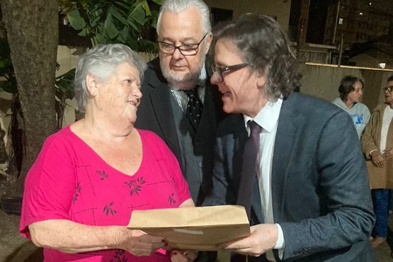 A imagem mostra três pessoas em destaque. À esquerda, há uma mulher idosa com cabelo grisalho, vestindo uma blusa rosa com detalhes florais escuros. Ela está segurando um objeto que parece ser um livro ou uma pasta. Ao lado dela, à direita, está um homem idoso de óculos, cabelo branco e barba, vestindo um terno escuro. À frente deles, há um homem de meia-idade com cabelo grisalho e óculos, vestindo um terno cinza e camisa branca. Ele também está segurando o mesmo objeto que a mulher. Ao fundo, há mais duas pessoas desfocadas, uma delas usando uma camiseta azul e a outra uma jaqueta marrom. A imagem parece ter sido tirada em um ambiente externo, com árvores ao fundo.