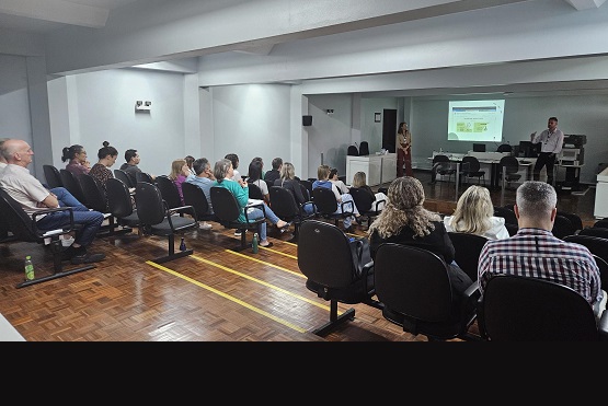 A imagem mostra uma sala de conferências ou treinamento, onde várias pessoas estão sentadas em cadeiras de auditório, todas voltadas para frente, assistindo a uma apresentação. A sala é simples, com paredes brancas e um piso de madeira envernizado. À frente, há um projetor exibindo um slide em uma tela grande. Dois apresentadores estão visíveis: um homem, de pé ao lado direito da sala, e uma mulher, que está de pé próxima ao centro, parecendo participar ativamente da apresentação. As pessoas estão organizadas em várias fileiras, prestando atenção ao que é apresentado. A atmosfera sugere um ambiente formal de palestra ou reunião profissional.