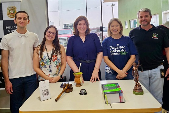 A imagem mostra cinco pessoas posando para uma foto em frente a uma mesa. Na parte de trás, há um homem de camiseta branca, uma mulher de óculos com uma blusa estampada, duas mulheres de blusa azul escura e um homem de blusa preta. Na mesa, há um suporte com um QR code, uma dama da justiça, uma chimarrão e alguns livros ou cadernos. Ao fundo, há um banner da Polícia Civil.