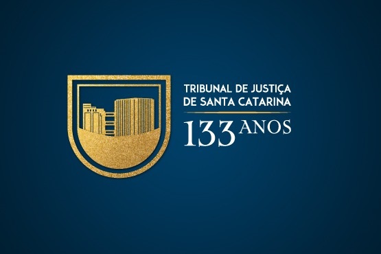 A imagem mostra um fundo azul escuro com um emblema dourado no lado esquerdo. O emblema parece ser um escudo com uma representação estilizada de um edifício ou colunas. À direita do emblema, está escrito em letras brancas "TRIBUNAL DE JUSTIÇA DE SANTA CATARINA" e abaixo, em números grandes, "133 ANOS".