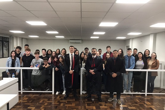 A imagem mostra um grupo de aproximadamente 30 pessoas em uma sala que parece ser um ambiente formal, possivelmente um tribunal ou uma sala de audiências. Na frente do grupo, há três homens em pé, usando trajes formais, incluindo togas pretas e gravatas vermelhas, sugerindo que podem ser advogados ou juízes. Atrás deles, há um grupo de jovens, possivelmente estudantes, que estão de pé em uma área cercada por uma grade branca. O piso é de madeira e o teto tem várias luminárias embutidas. A atmosfera parece ser de um evento educacional ou uma visita guiada.