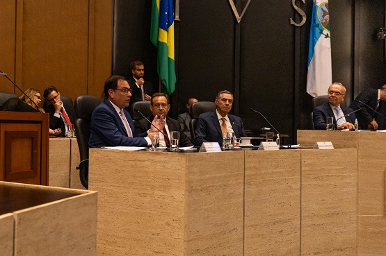 A imagem retrata um evento formal em um ambiente institucional, provavelmente um debate ou conferência. Cinco indivíduos estão sentados atrás de uma longa mesa em um auditório. Eles estão em frente a microfones, indicando que estão envolvidos em uma discussão ou apresentação. O homem no centro, que parece estar falando, está vestido com um terno escuro e óculos. Os outros participantes, também vestidos formalmente, estão atentos, com um deles fazendo uma anotação. O cenário inclui bandeiras do Brasil e de um estado ou organização, e a sala tem um aspecto austero e formal, com acabamentos em madeira e pedra.