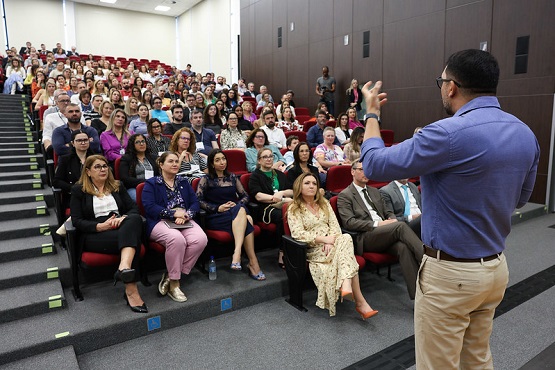 A imagem mostra uma cena em uma sala de conferências moderna. Um homem de pé, vestindo camisa azul e calça bege, está apresentando ou falando para uma audiência atenta. Ele está de costas para a câmera, gesticulando com as mãos enquanto fala. O público, composto por cerca de cinquenta pessoas sentadas em fileiras de cadeiras vermelhas, parece engajado, com alguns membros olhando diretamente para o palestrante e outros anotando. No primeiro plano, há três filas de cadeiras com ocupantes que vestem trajes formais e casuais, demonstrando um ambiente profissional e diversificado.