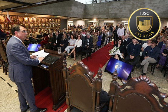 A imagem mostra uma cerimônia formal. À esquerda, um homem de terno está em pé atrás de um púlpito, falando para a audiência. Ele está segurando papéis e há um copo de água ao lado dele. No centro, há um corredor coberto por um tapete vermelho, e as cadeiras estão dispostas em fileiras de cada lado. A audiência é composta por homens e mulheres, muitos deles vestidos formalmente. Ao fundo, há uma parede com retratos emoldurados. O ambiente é espaçoso, com um teto alto e paredes de concreto aparente.