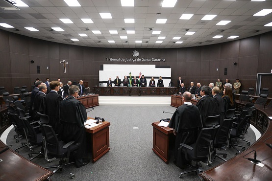 A imagem mostra uma sala de tribunal ampla e moderna, com um grande número de pessoas presentes. No centro, há uma bancada elevada onde estão sentados vários juízes, todos vestidos com togas pretas. Atrás deles, há uma parede com o texto "Tribunal de Justiça de Santa Catarina". A sala é bem iluminada, com um teto repleto de luminárias embutidas. À esquerda, há um crucifixo na parede. As pessoas na sala estão em pé, possivelmente participando de uma cerimônia ou sessão formal. As cadeiras são de cor preta e estão dispostas em semicírculo ao redor da bancada central.
