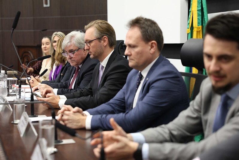 A imagem mostra uma mesa de conferência com várias pessoas sentadas lado a lado. Há seis pessoas visíveis na imagem, todas vestidas formalmente com ternos e gravatas. A maioria delas está olhando para a frente ou para baixo, possivelmente para documentos ou dispositivos eletrônicos. No fundo, há uma bandeira do Brasil. A mesa está equipada com microfones e copos de água. O ambiente parece ser uma sala de reuniões ou conferências, com um painel de madeira escura ao fundo.