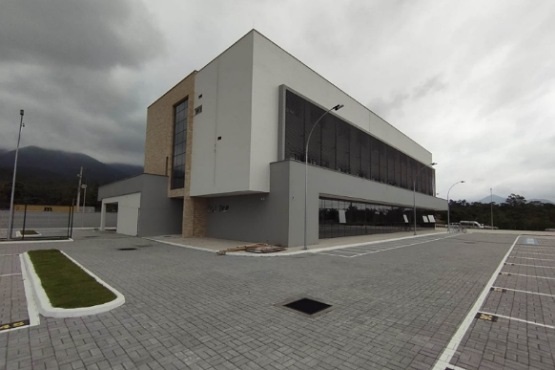A imagem mostra um edifício moderno de dois andares, com um design geométrico e linhas retas. A fachada é predominantemente cinza, com detalhes em vidro e uma seção revestida com tijolos aparentes. O prédio está localizado em um amplo estacionamento pavimentado com blocos de concreto, que está vazio. Há postes de iluminação ao redor e algumas áreas verdes pequenas com grama. O céu está nublado, sugerindo um dia cinzento e possivelmente chuvoso. Ao fundo, é possível ver uma cadeia de montanhas coberta por nuvens.