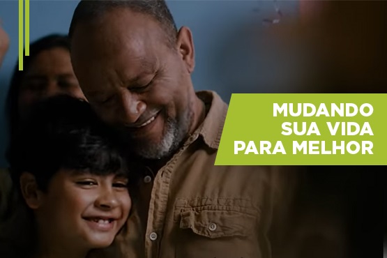 A imagem mostra um homem mais velho, com barba grisalha, sorrindo e abraçando uma criança que também está sorrindo. O fundo é desfocado, mas parece ser de cor azul. No lado direito da imagem, há um retângulo verde com o texto em branco que diz: "MUDANDO SUA VIDA PARA MELHOR".