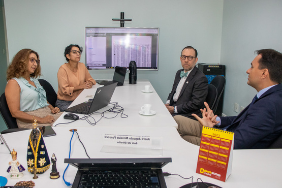 A imagem retrata uma reunião de negócios em um escritório. Quatro pessoas estão sentadas ao redor de uma mesa retangular, três homens e uma mulher, todos focados em uma discussão. A mulher e um dos homens estão usando laptops. No centro da mesa, há uma tela de computador grande exibindo planilhas ou dados, indicando que a reunião pode envolver análise financeira ou de dados. Cada pessoa exibe uma postura profissional; o homem à direita gesticula enquanto fala, indicando que ele pode estar explicando um ponto ou liderando a discussão. Na mesa, há xícaras de café e objetos pessoais como livros e decorações religiosas, incluindo uma imagem de Nossa Senhora e um rosário, adicionando um toque pessoal ao ambiente de trabalho formal. O cenário transmite uma atmosfera de trabalho colaborativo e sério, típico de um ambiente corporativo ou acadêmico focado em resultados e discussões detalhadas.