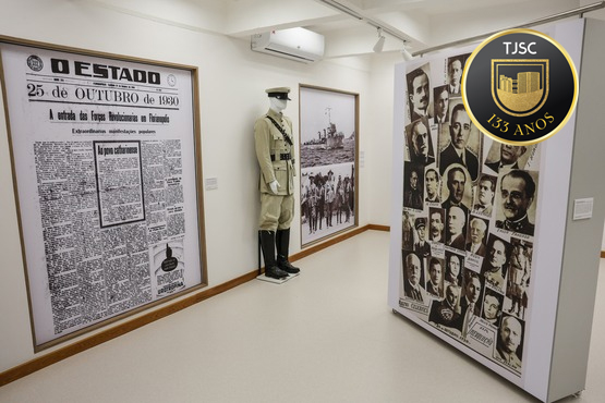 A imagem mostra uma exposição em um museu ou espaço de memória. À esquerda, há um manequim vestido com um uniforme militar branco, possivelmente de um almirante, dado o estilo do uniforme e a presença de um quepe. À direita, dois painéis exibem informações e imagens históricas: o primeiro é uma reprodução de uma primeira página do jornal "O Estado" datada de 25 de outubro de 1980, discutindo um evento militar. O segundo painel mostra fotografias de várias figuras históricas em uniforme. O ambiente é bem iluminado e projetado para a educação histórica, provavelmente focando em eventos militares ou figuras importantes de um contexto específico.