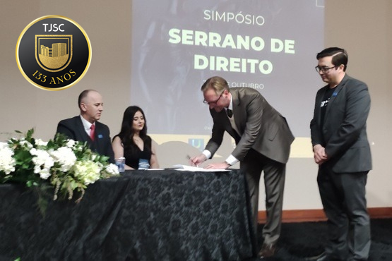 A imagem mostra quatro pessoas em um evento acadêmico. Um homem à direita, vestido com terno cinza, está se inclinando para assinar um documento sobre uma mesa coberta com uma toalha preta. À sua esquerda, há três pessoas observando a assinatura: um homem careca vestindo terno vermelho, uma mulher de cabelo preto e blusa azul, e um homem de óculos e terno preto. Ao fundo, um slide projeta o título "SIMPÓSIO SERRANO DE DIREITO". O ambiente é formal, típico de conferências ou cerimônias acadêmicas.
