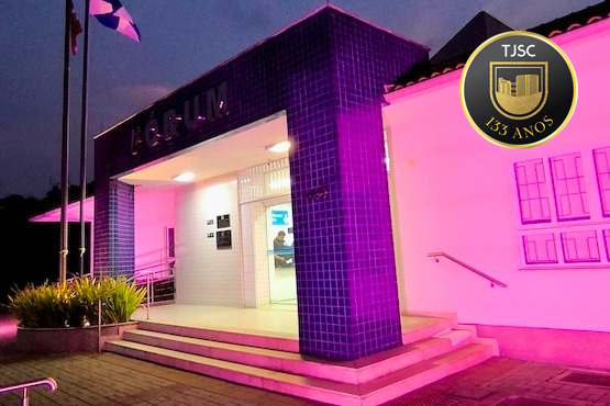 A imagem mostra a entrada de um edifício, iluminado com luzes rosa e roxas que criam um ambiente vibrante e moderno. A fachada é marcada por azulejos roxos e um letreiro que diz "FORUM", indicando que o edifício é um fórum ou tribunal. Há uma bandeira ao lado da entrada, embora o país ou estado representado pela bandeira não seja visível. A entrada principal é acessada por uma escada com alguns degraus, e as paredes externas são iluminadas para destacar a arquitetura. A iluminação noturna e o design único sugerem que o local pode ter importância cívica ou judicial, com um toque estético contemporâneo para atrair a atenção pública ou celebrar algum evento especial.