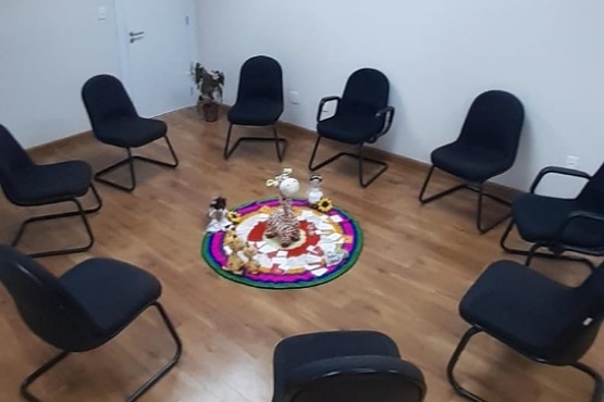A imagem mostra uma sala com piso de madeira e paredes brancas. Há nove cadeiras pretas dispostas em círculo no centro da sala. No meio do círculo, há um tapete colorido com vários objetos em cima, incluindo bonecos, flores e cartões. Ao fundo, há uma porta branca e uma planta em um vaso no canto da sala. A disposição sugere um ambiente preparado para uma reunião ou atividade em grupo.
