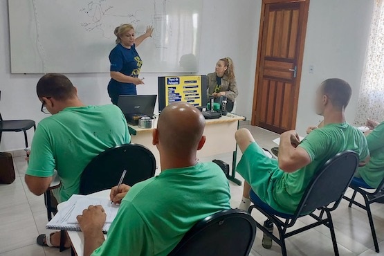 A imagem mostra uma sala de aula com seis pessoas. Quatro delas estão sentadas em cadeiras, aparentemente assistindo a uma apresentação ou aula. Elas estão vestindo camisetas verdes. Uma mulher está em pé na frente da sala, ao lado de um quadro branco, apontando para algo no quadro. Ela está vestindo uma camiseta azul. Ao lado dela, uma outra mulher está sentada atrás de uma mesa, onde há um laptop, alguns potes e um cartaz com texto. A sala tem paredes brancas e uma porta de madeira. Há também uma cortina clara cobrindo uma janela à direita.