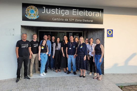 A imagem mostra um grupo de 14 pessoas posando para uma foto em frente a um prédio identificado como "Justiça Eleitoral, Cartório da 57ª Zona Eleitoral". As pessoas estão em pé, lado a lado, sorrindo para a câmera. Elas estão vestidas de maneira casual e formal, com algumas usando crachás. Acima da entrada do prédio, há uma placa com o brasão da Justiça Eleitoral e o nome do cartório. Ao lado direito da entrada, há um pequeno cartaz com o símbolo da Justiça Eleitoral. O chão é de ladrilhos claros e a fachada do prédio é pintada de branco.