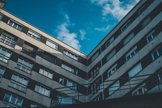 A imagem mostra um edifício residencial moderno, de média altura, com várias janelas e varandas. O edifício é de cor predominantemente branca com detalhes em preto. Há uma estrutura de escada externa visível, que parece ser feita de metal, dando um aspecto utilitário ao prédio. O céu acima está claro, com algumas nuvens, o que sugere que a foto foi tirada em um dia ensolarado. A perspectiva da foto é ligeiramente inclinada para cima, focando na simetria e linhas arquitetônicas do edifício contra o céu azul.
