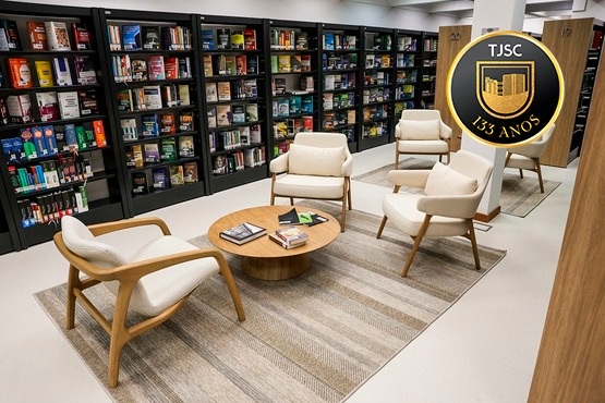 A imagem mostra o interior de uma biblioteca moderna e elegante. A área é bem iluminada e possui prateleiras escuras cheias de livros que cobrem as paredes ao fundo. No centro do espaço, há um conjunto de móveis composto por quatro poltronas brancas de design contemporâneo, dispostas ao redor de uma mesa de centro redonda de madeira. Sobre o tapete cinza, que cobre parte do piso,há uma mesa de centro onde estão dispostos alguns livros e revistas. No canto superior direito da imagem, há um emblema com o texto "TJSC 133 ANOS". A atmosfera é acolhedora, ideal para leitura e discussões tranquilas.